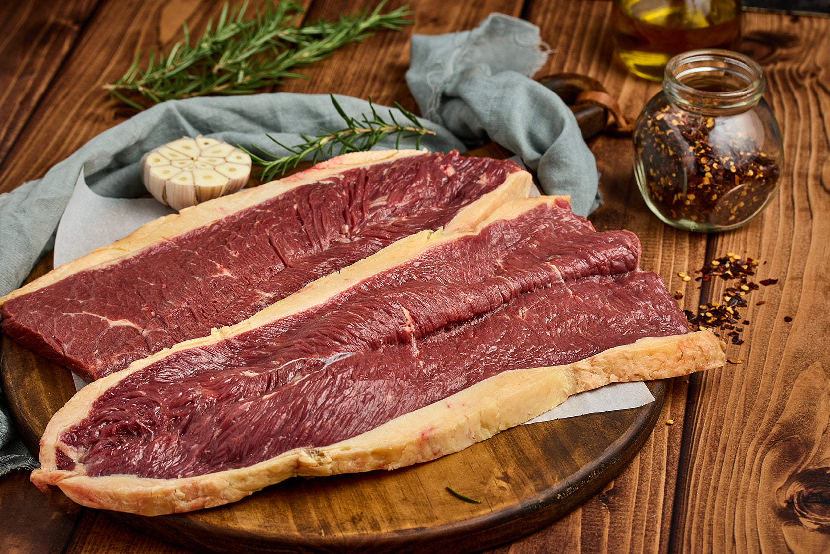 Imagen de una carne sobre una tabla de madera, acompañada de especias, ajo y romero